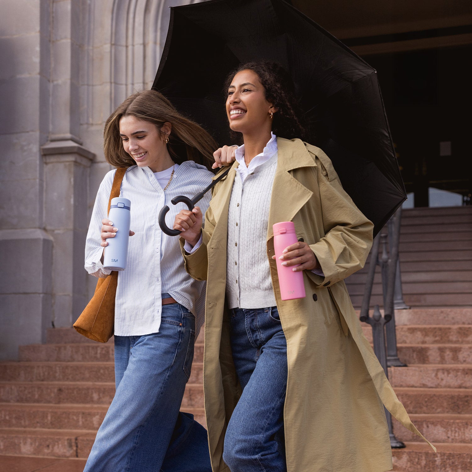 Two women walking down set of stairs outside a building. Each is holding a Sidekick coffee tumbler. One is also holding a purse and the other an umbrella.