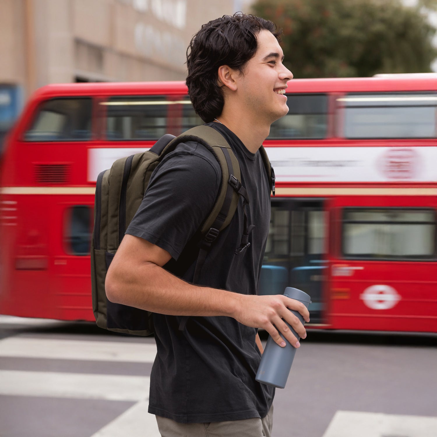 Man walking outside carrying a backpack and a Sidekick coffee tumbler. Bus is driving by in the background.