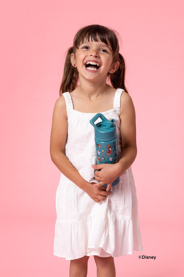 Young girl in white dress holding a teal Disney Moana water bottle against a pink background, smiling at camera in studio setting.