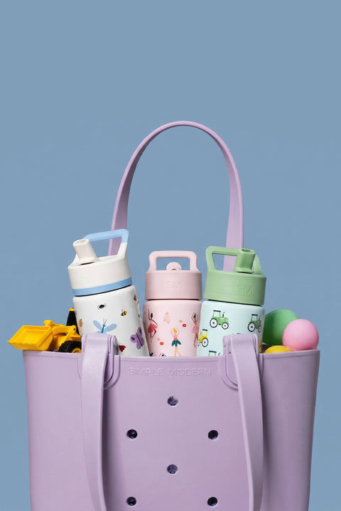 Lavender-pink Simple Modern tote bag filled with three pastel insulated tumblers featuring bee and floral designs, colorful balls, and yellow accessories on a blue background.