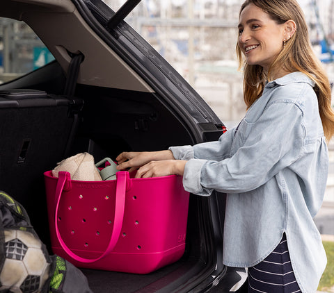 Woman loading a hot pink Simple Modern Getaway Bag into a car trunk in an urban setting, with a soccer ball nearby and city buildings visible in the background.