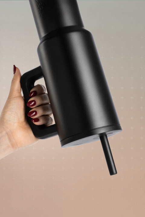Black matte drinkware displayed against a warm beige and cream gradient background, held by a person with peachy-beige skin tone.