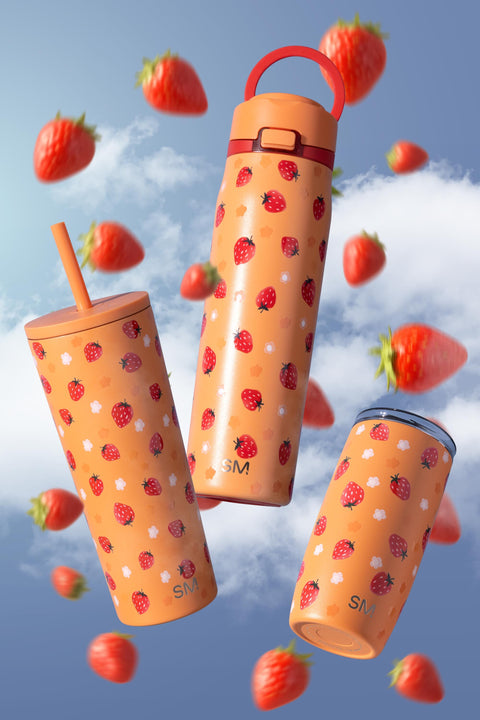 Three peachy-orange drinkware pieces surrounded by fresh red strawberries with green leaves against a soft blue sky with white clouds.