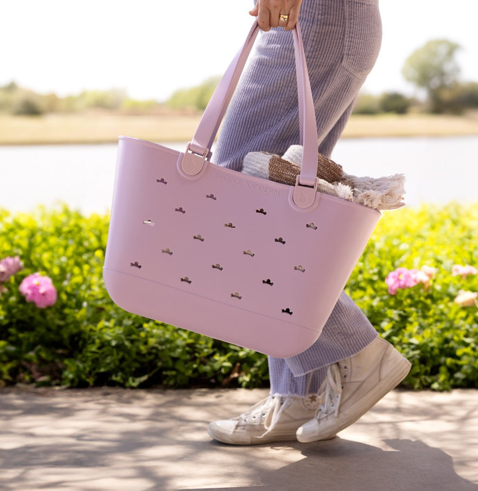 A woman walks past a flowering hedge beside a lake, carrying a pale pink Simple Modern tote bag with Mickey-shaped cutouts.