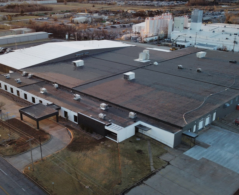 Industrial rooftop with dark gray and charcoal flat surfaces, white HVAC units and ductwork, and asphalt patches showing domestic manufacturing facility infrastructure.