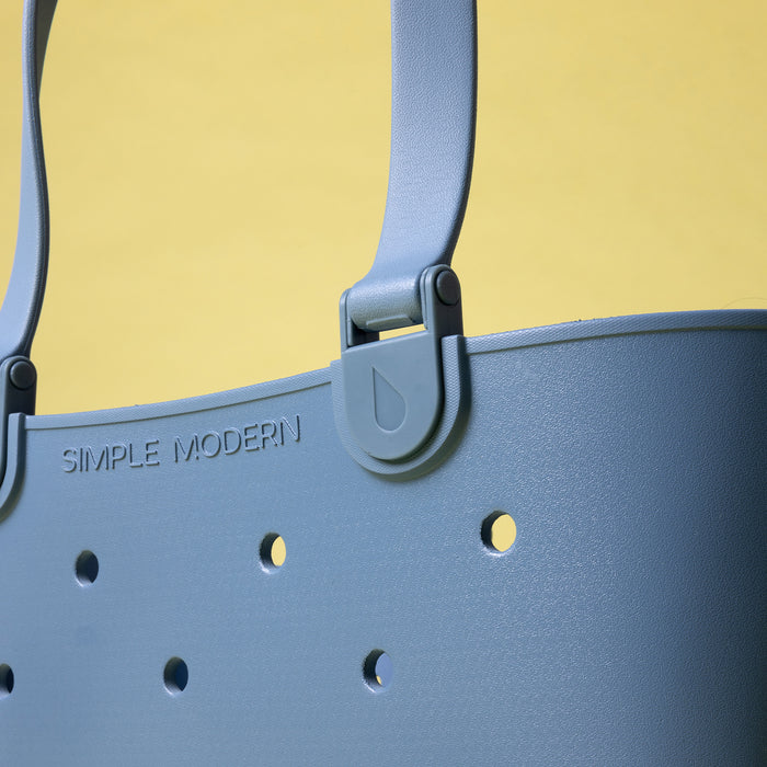Close-up of a slate blue Getaway Bag tote showing its reinforced handle clip, embossed Simple Modern logo and drainage holes.