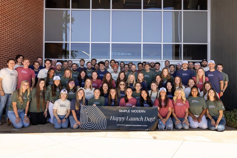 Large group of approximately 70 team members arranged in rows in front of brick building with white-framed windows, wearing cream-colored branded apparel.