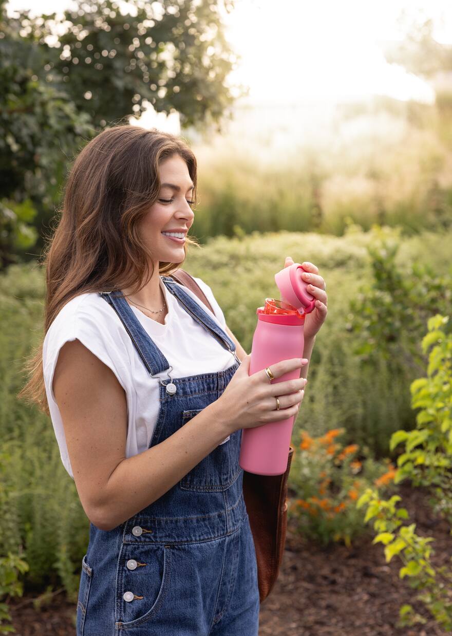 Woman in denim overalls opening a pink insulated water bottle in a sunlit garden