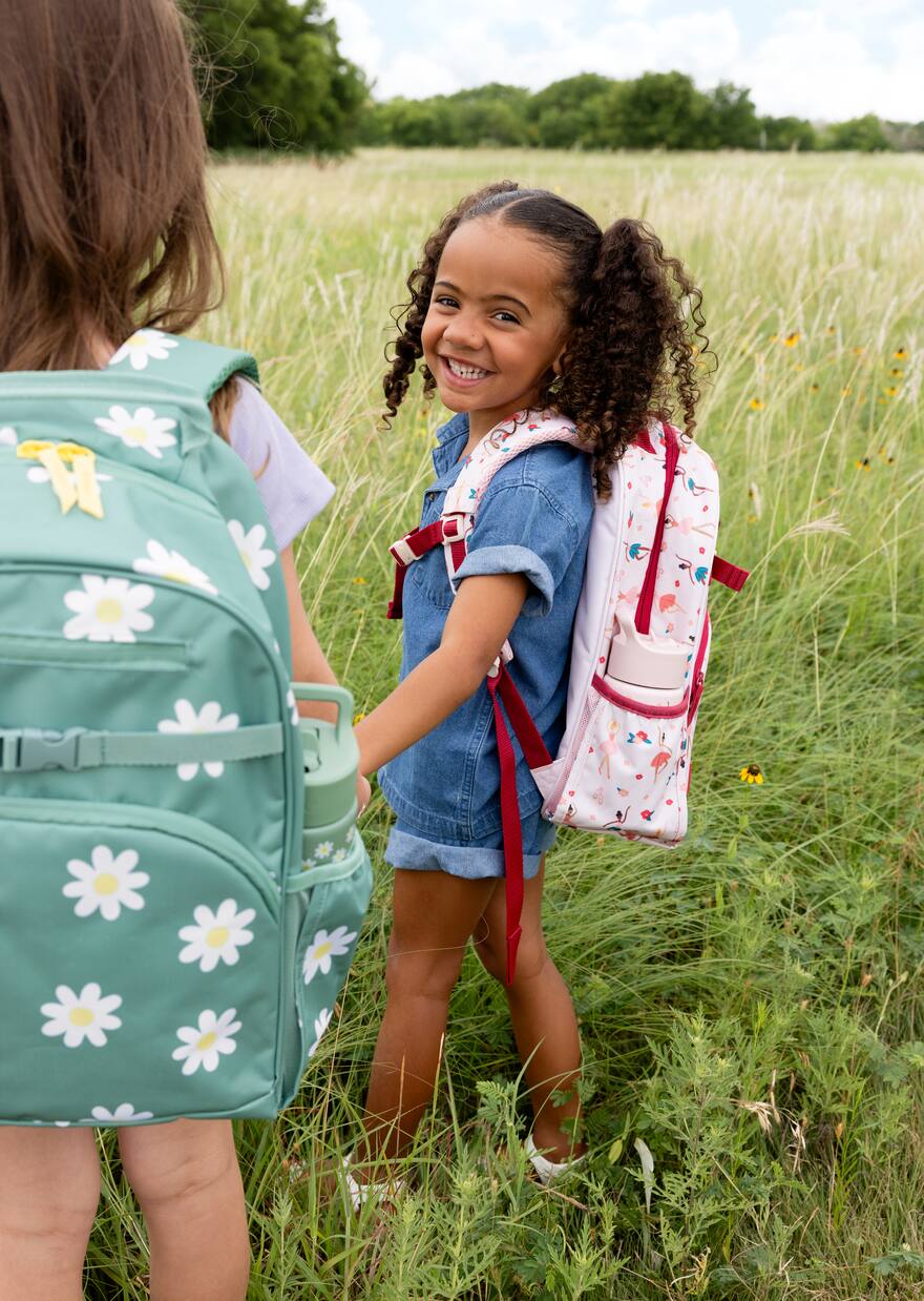 Two children wearing floral backpacks smiling at each other in a grassy field